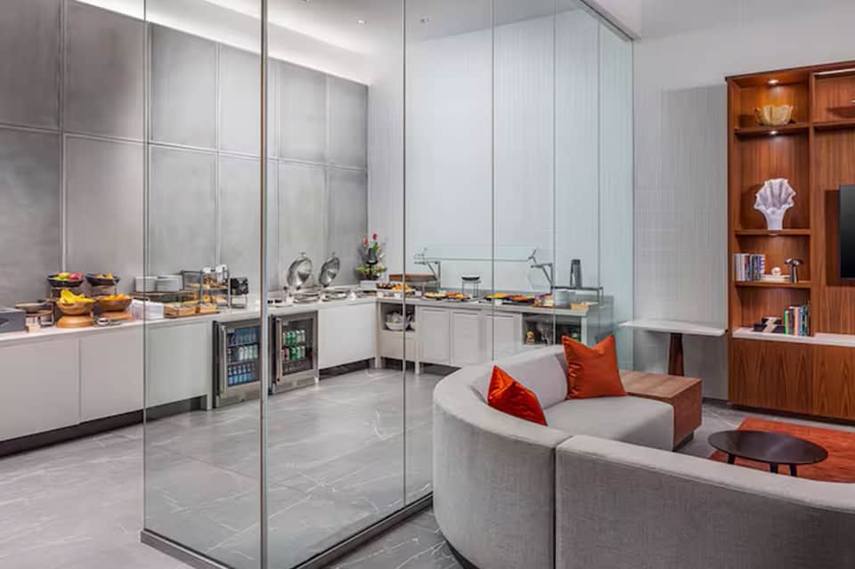 Contemporary hotel lounge with curved seating, kitchenette, glass partitions, and wood shelving with accent lighting