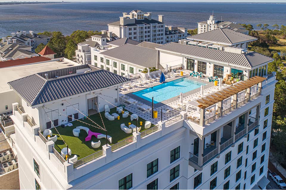 Aerial view of waterfront resort with pool, rooftop lounge area, green space, and ocean backdrop