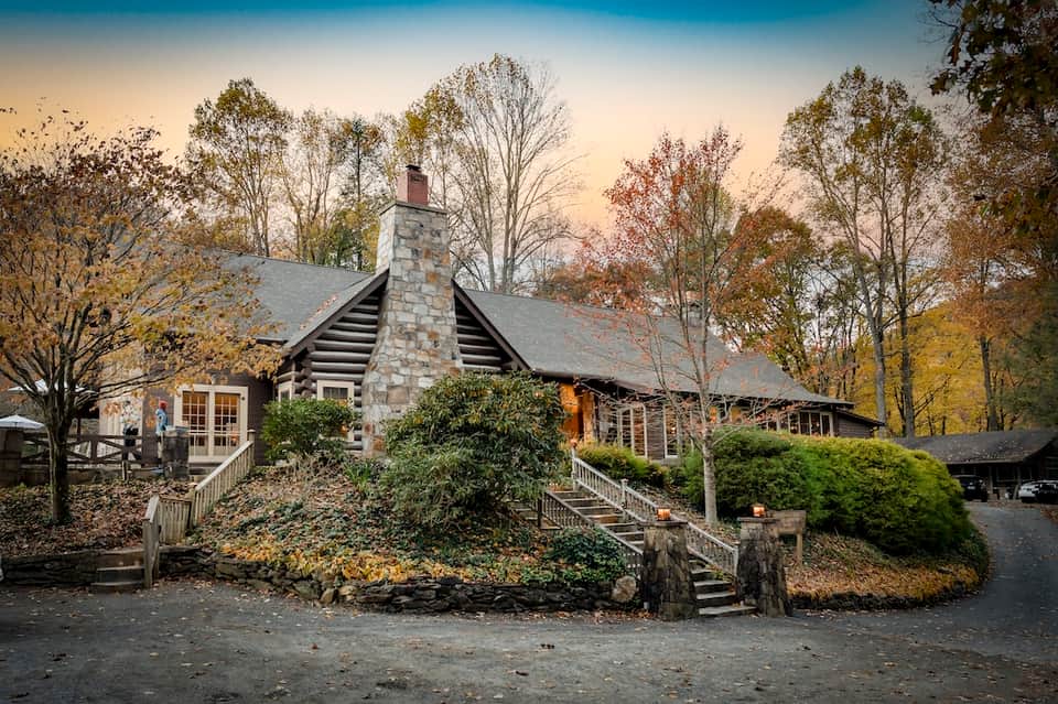 Rustic log cabin with stone chimney surrounded by autumn trees and wooden bridges
