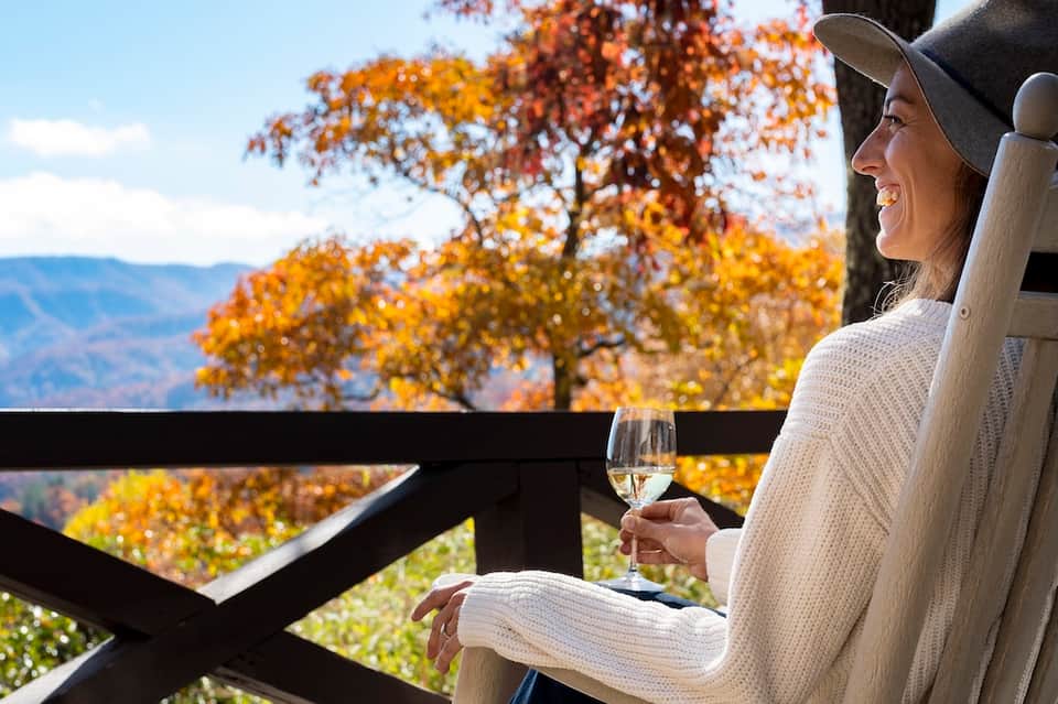 Woman in white sweater and hat holding wine glass on autumn balcony overlooking fall foliage and mountains