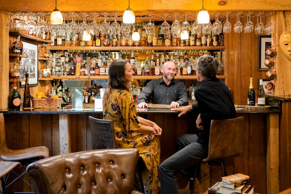 Wooden bar interior with bartender serving guests, well-stocked shelving, and ambient pendant lighting
