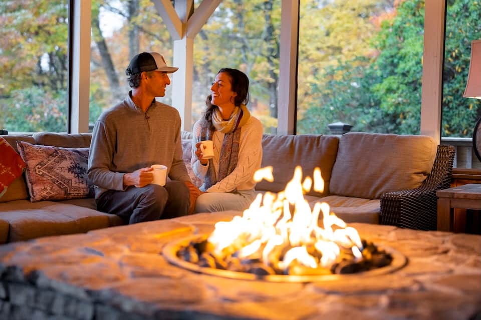 Couple sitting by outdoor fire pit on brown wicker furniture holding coffee cups