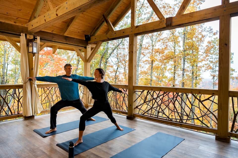Couple practicing yoga on mats in a glass-enclosed pavilion overlooking autumn foliage