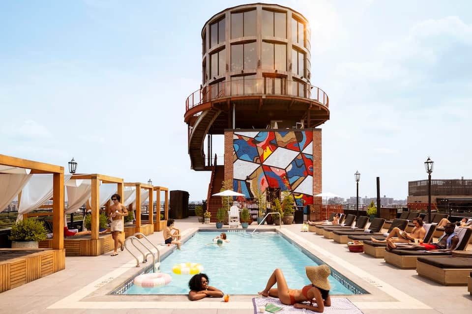 Rooftop pool with water tower sculpture, loungers, cabanas, and umbrellas overlooking city skyline