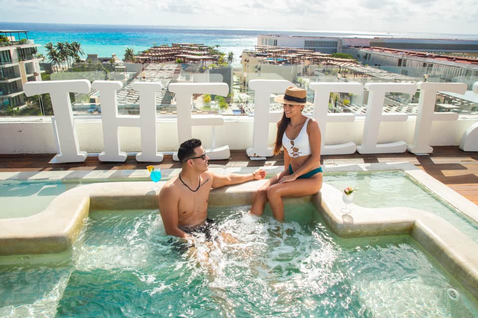Man and woman in hot tub at rooftop pool overlooking beachfront resort with turquoise water