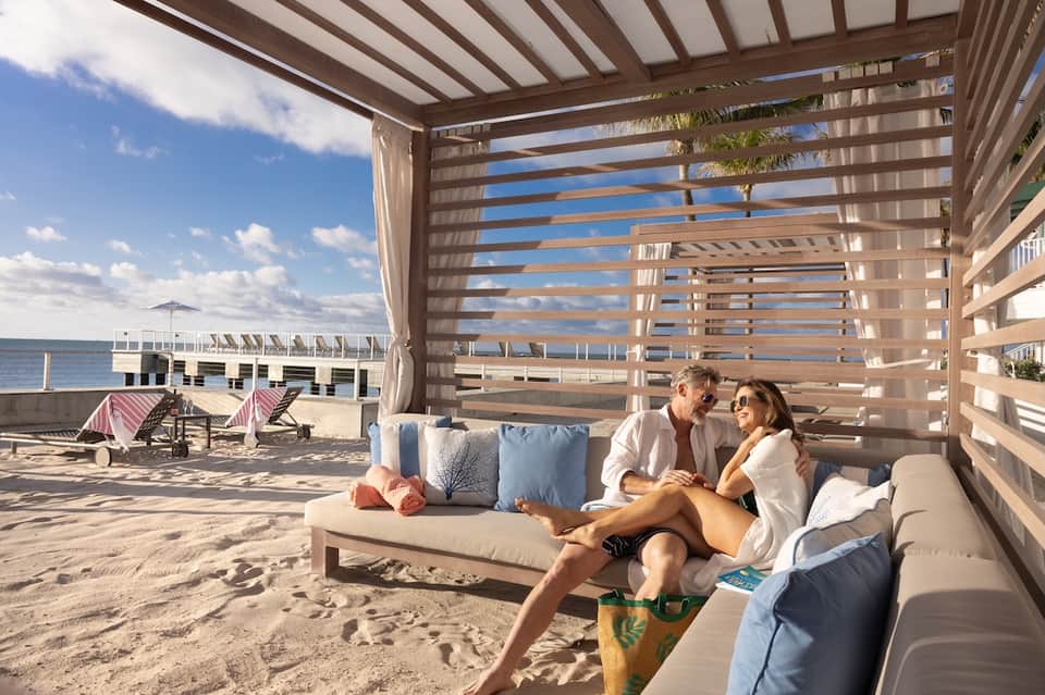 Beachfront cabana with couple relaxing on cream sofa under wooden pergola with ocean and sky views