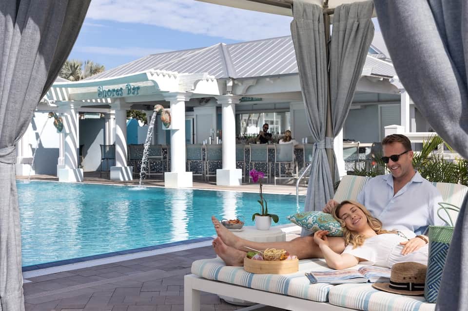 Couple relaxing on poolside cabana bed overlooking resort pool and Shores Bar facility