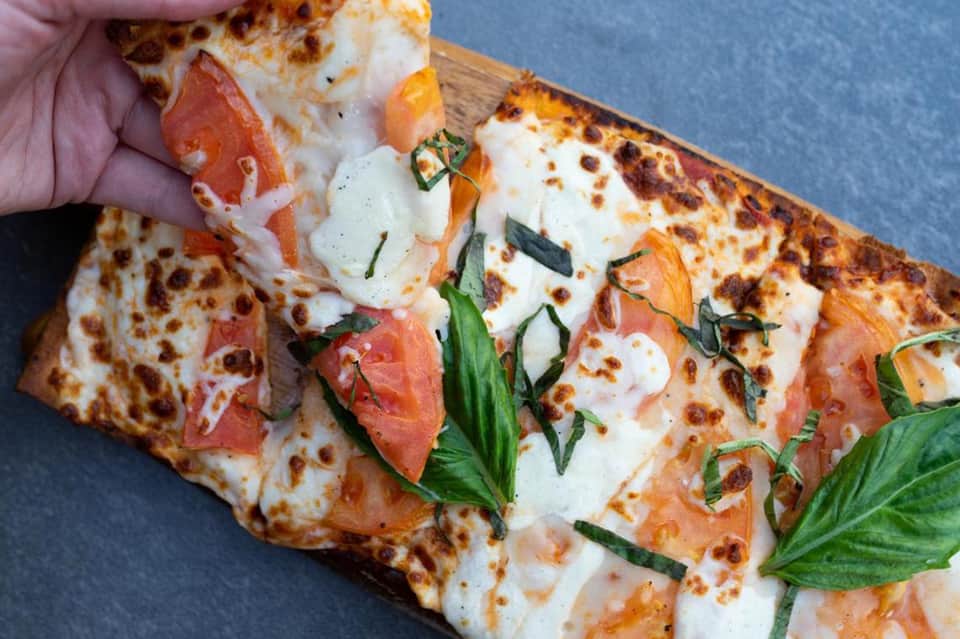 Slice of pizza with melted cheese, prosciutto, fresh basil leaves being held against blue background