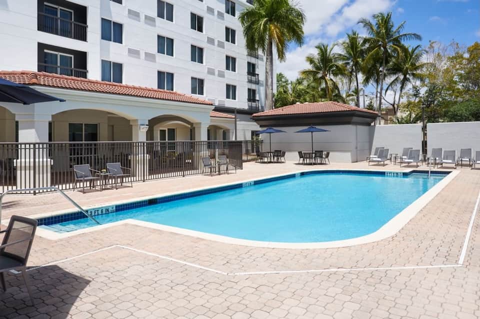 Modern hotel pool with white lounge chairs, umbrellas, metal railings, and multi-story white building