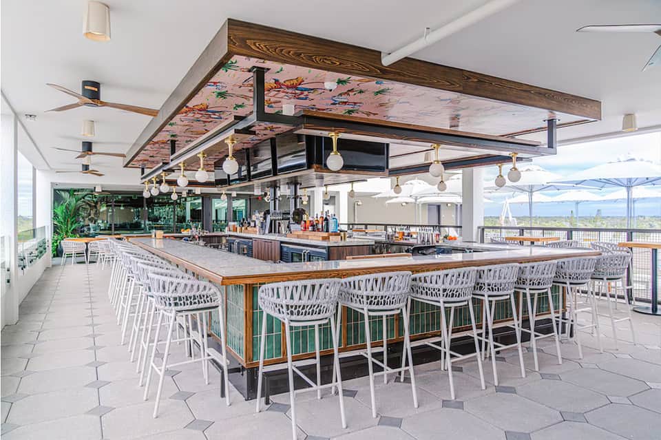 Modern rooftop bar with waterfront views, wood-beam ceiling, pendant lights, and white bar stools