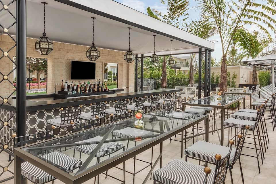 Upscale outdoor patio bar with black modern design, hanging pendant lights, geometric patterned chairs, and landscaping