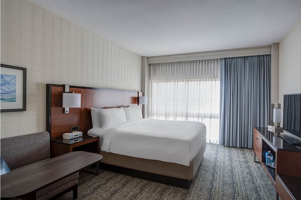 Hotel bedroom with queen bed, wood headboard, modern furnishings, and ocean-view balcony