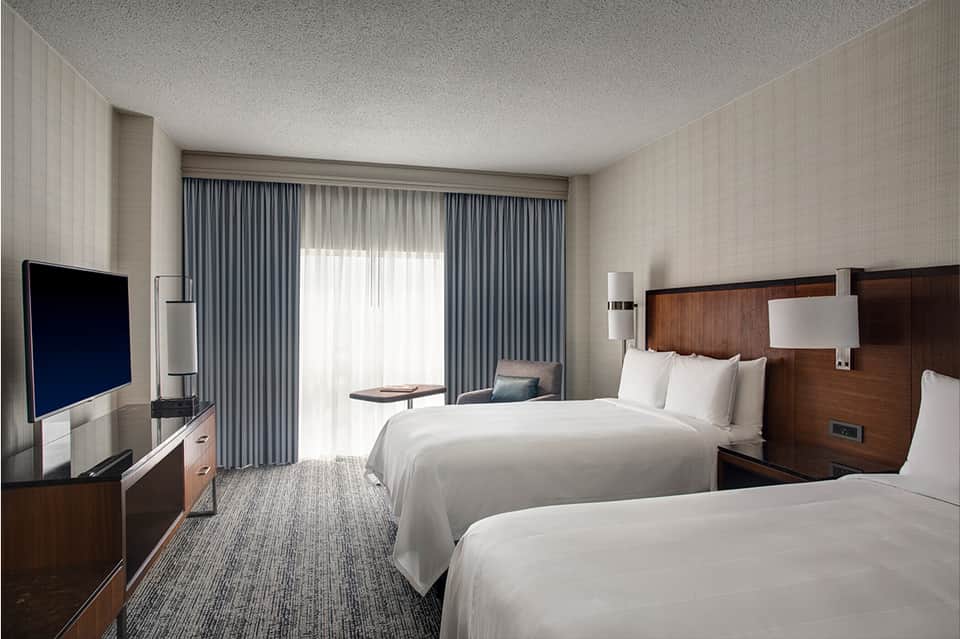 Modern hotel room with two queen beds, flat-screen TV, balcony doors, and contemporary neutral decor