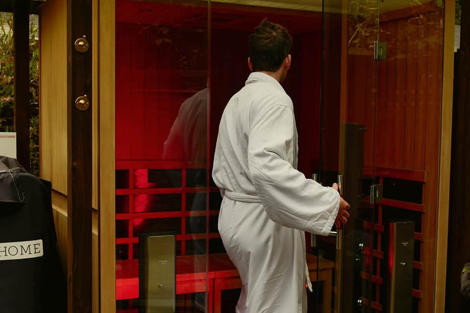 Person in white robe entering red infrared sauna room with glass doors