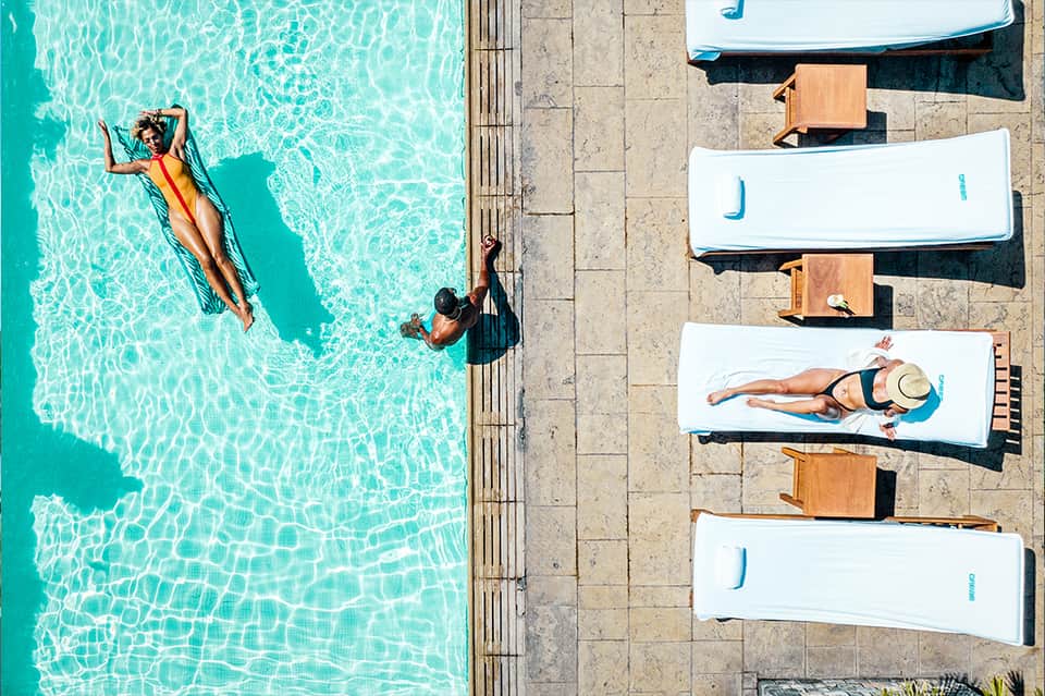 Aerial view of resort pool with swimmers, lounge beds with cushions, and concrete deck areas
