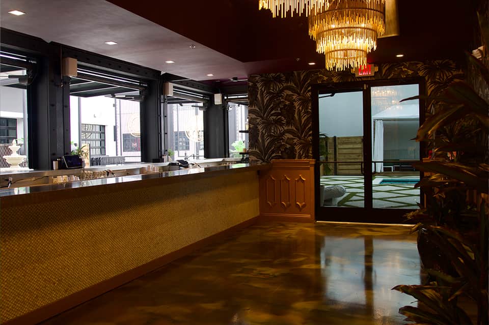 Modern hotel lobby with gold chandelier, dark floral wallpaper, polished concrete floor, and sleek reception desk.