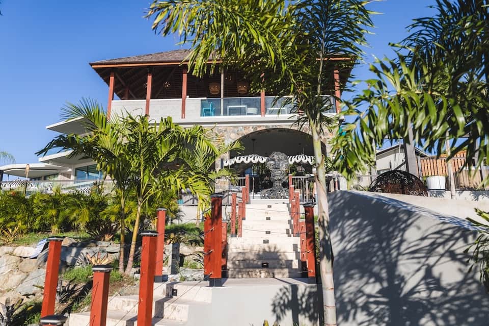 Beach resort entrance with red wooden gate, palm trees, and white umbrellas visible beyond