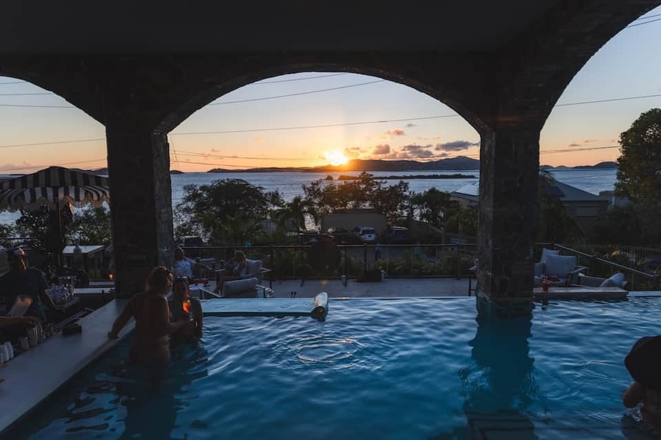 Infinity pool overlooking bay at sunset with concrete pillars and guests swimming