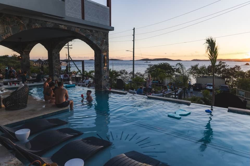 Infinity pool overlooking bay at dusk with guests, stone archway, and ocean sunset views