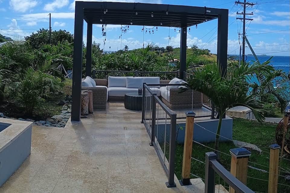 Oceanside terrace with black pergola, wicker seating, hanging lights, and tropical coastal views