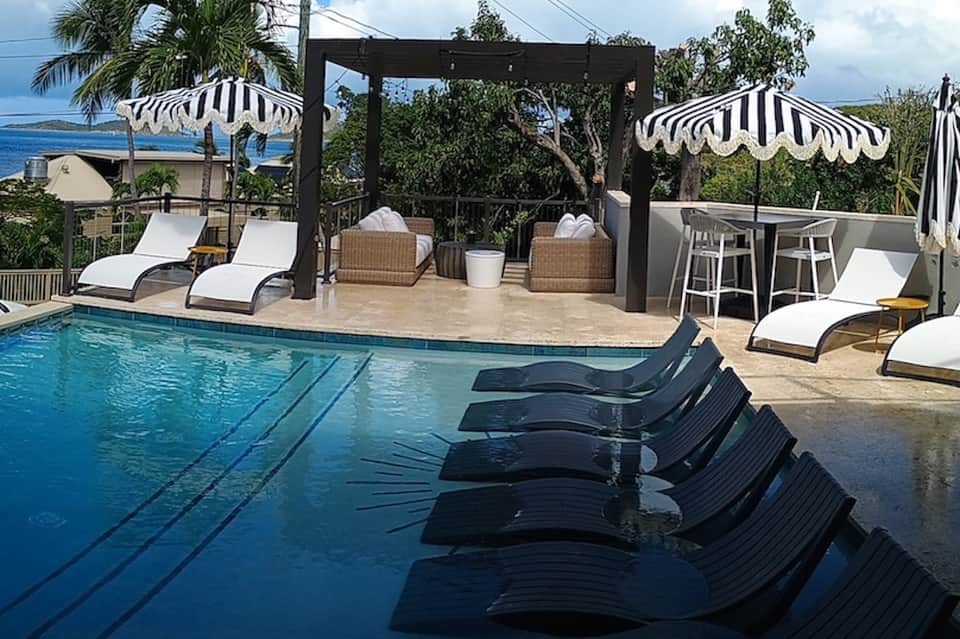 Resort pool area with black loungers, striped umbrellas, bar seating, and ocean view