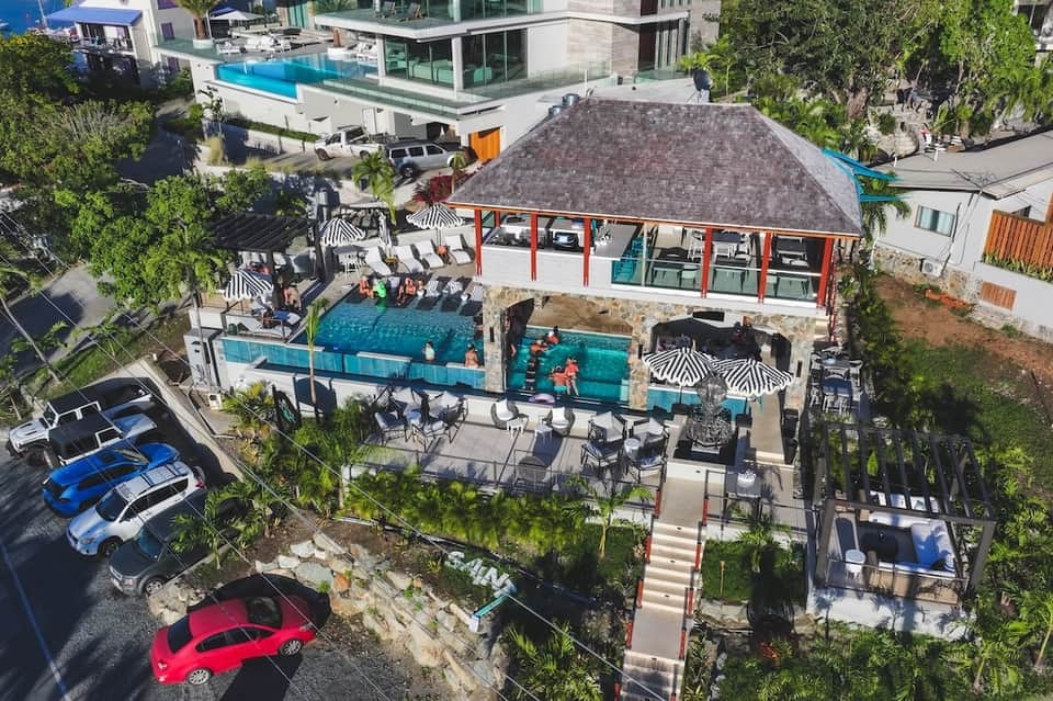 Aerial view of contemporary pool area with shade structures, multiple pools, lounging spaces, and surrounding homes
