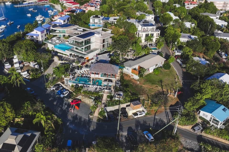 Aerial view of waterfront homes with pools, modern architecture, boats, lush green trees, and blue water