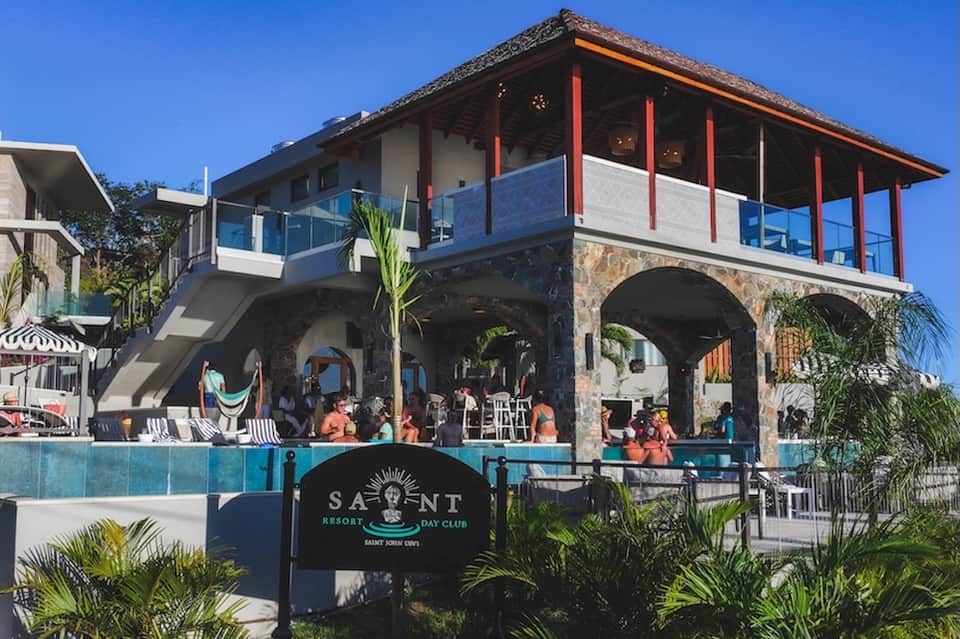Beachfront day club with multi-level architecture, pool, palm trees, and covered lounge areas