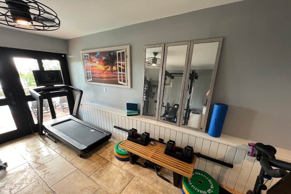 Home gym with black treadmill, mirrored wall, dumbbells on wooden bench, and bright natural light from French doors