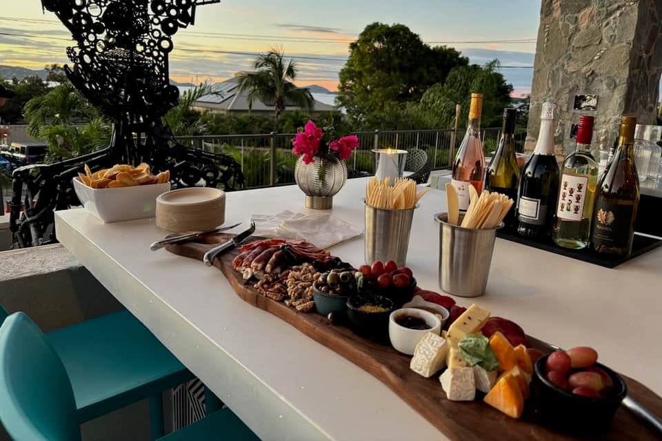 Sunset view from resort bar with charcuterie spread, wine bottles, and waterfront horizon