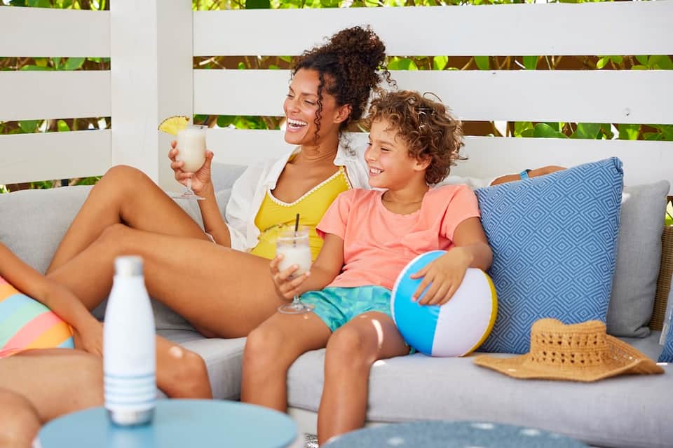 Woman and child enjoying drinks on gray outdoor sofa with colorful beach ball and accessories