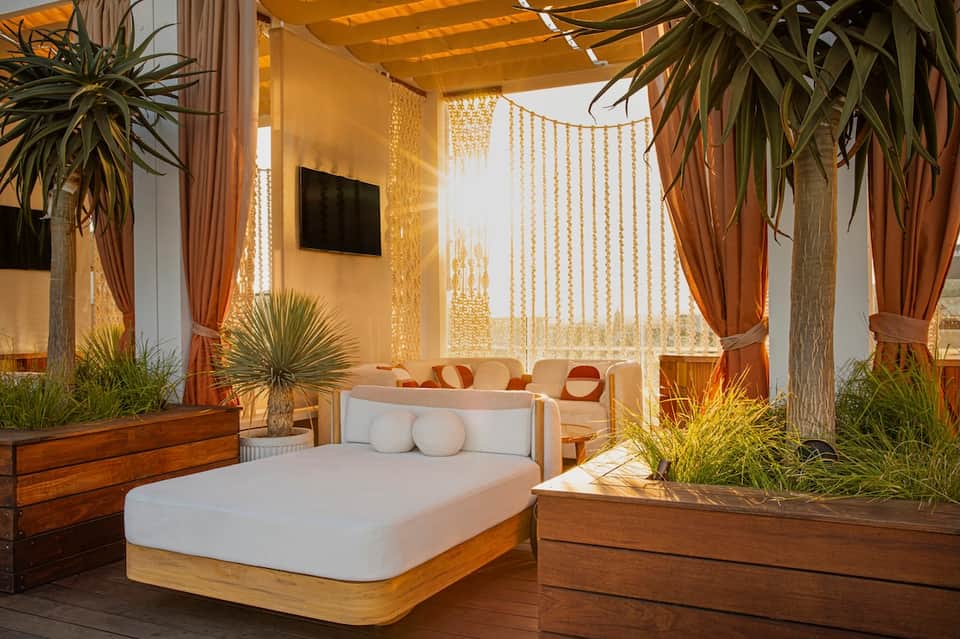 Contemporary bedroom with wooden bed frame, white linens, potted plants, and draped curtains with ocean view