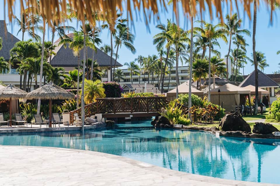 Resort pool with wooden bridge, palm trees, and tiki umbrellas under blue sky