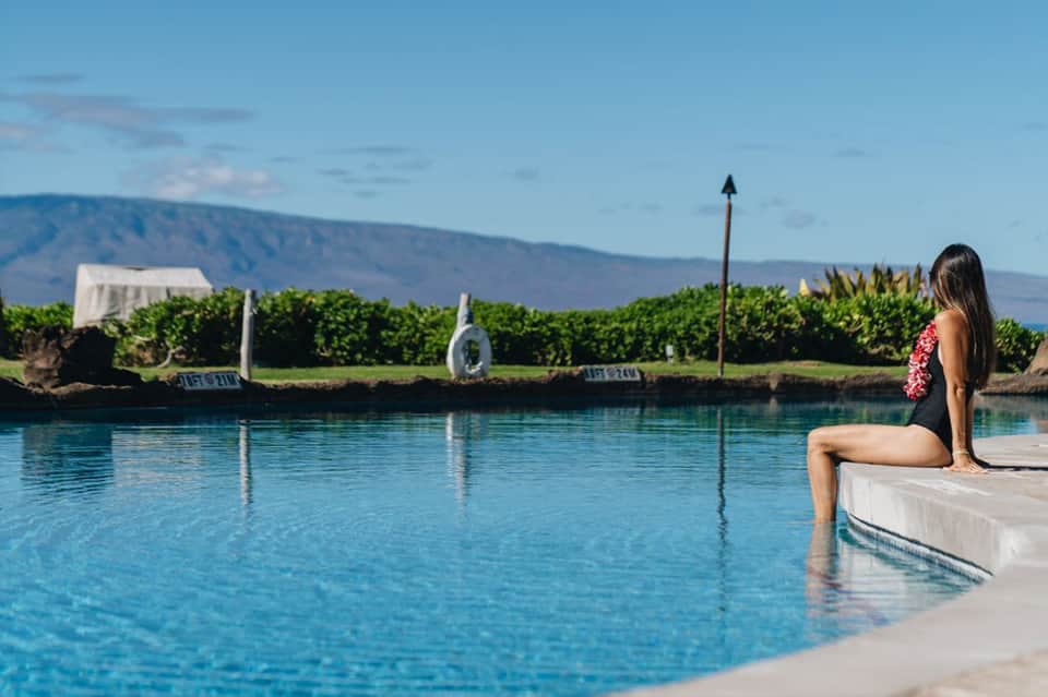 Woman sitting at edge of crystal-blue pool overlooking scenic waterfront landscape with mountains