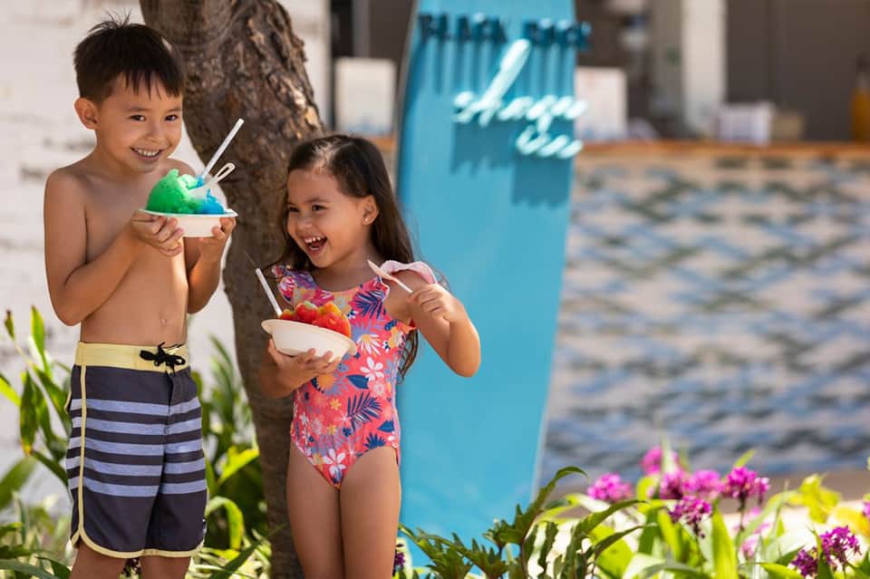 Two children enjoying colorful shaved ice drinks on beach near palm tree and flowers