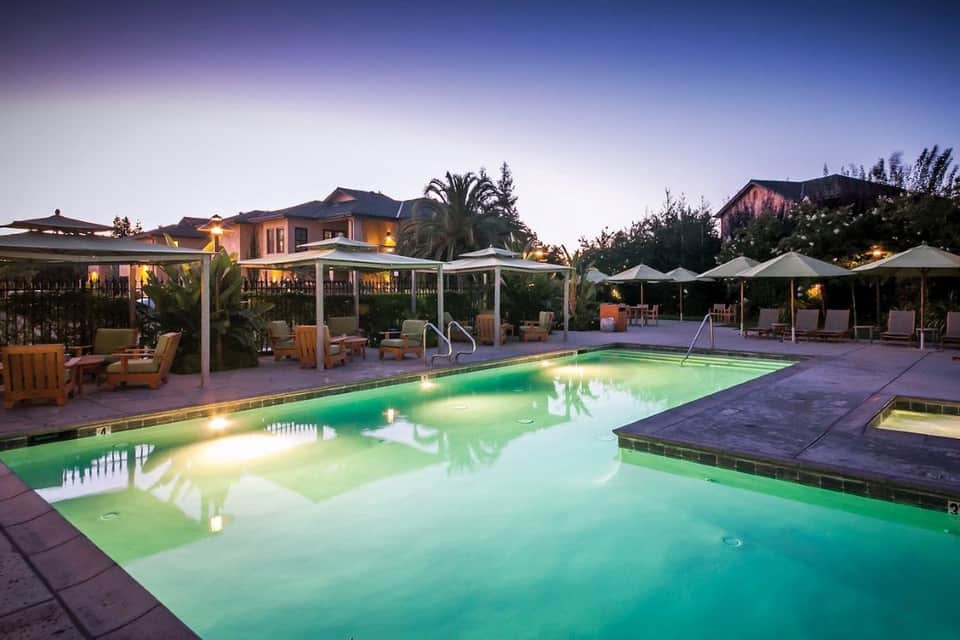 Illuminated resort pool at dusk with white pergolas, lounge chairs, palm trees, and multi-story buildings
