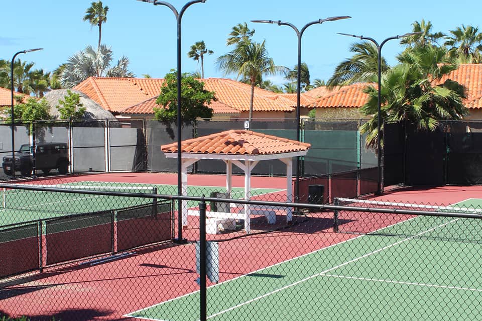 Tennis courts with orange clay surface, covered seating area, palm trees, clear blue sky, well-maintained facility