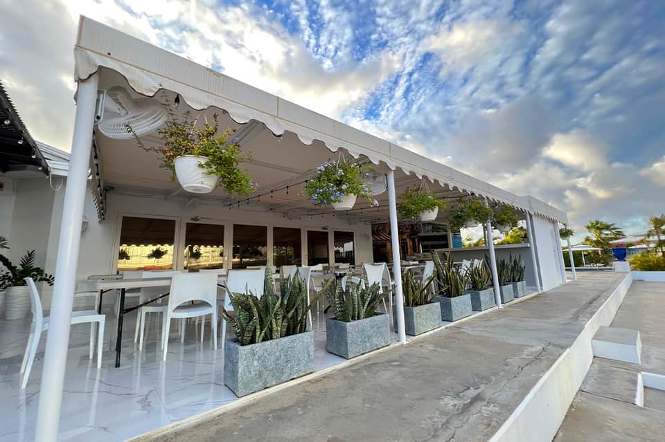 Modern white pergola restaurant with retractable awning, bar seating, potted plants, and cloud sky