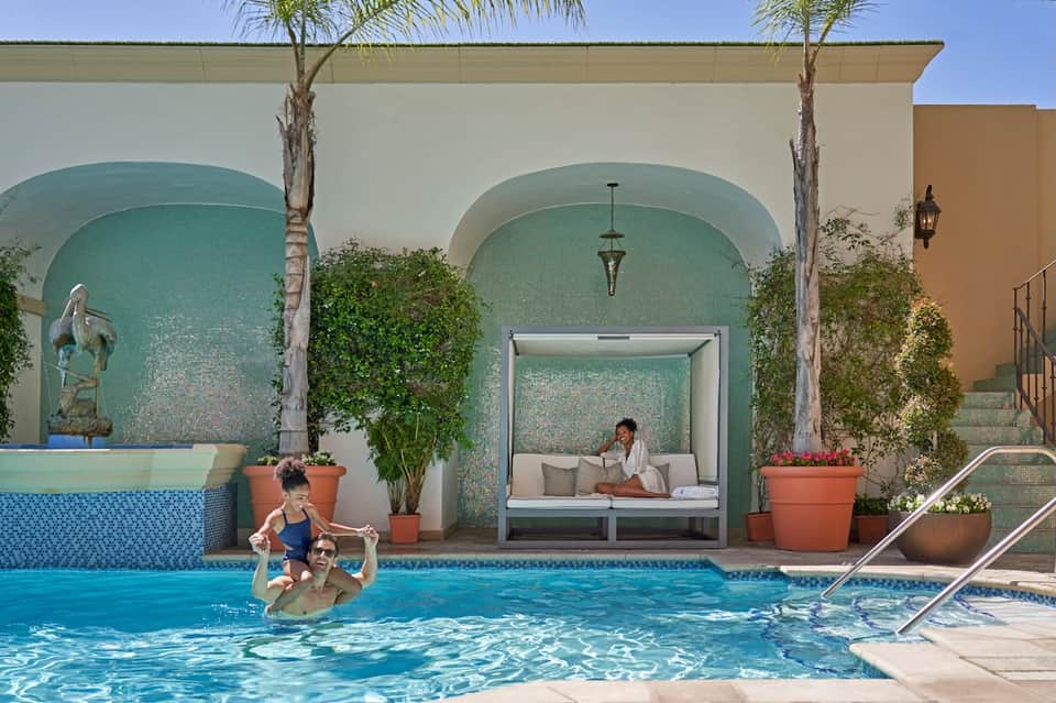 Resort pool area with turquoise water, arched teal tile walls, palm trees, and lounge pavilion