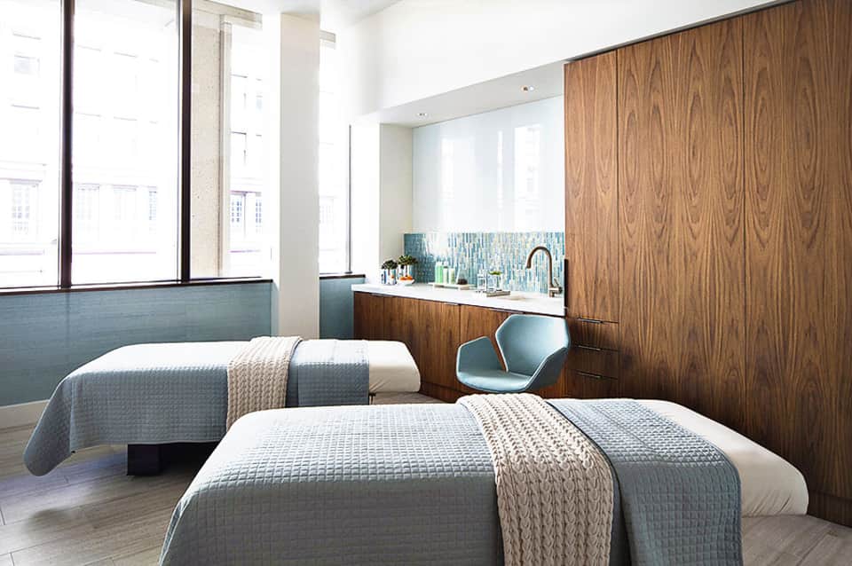 Modern hotel bedroom with two beds, wood paneling, large windows, and integrated bathroom with sink visible