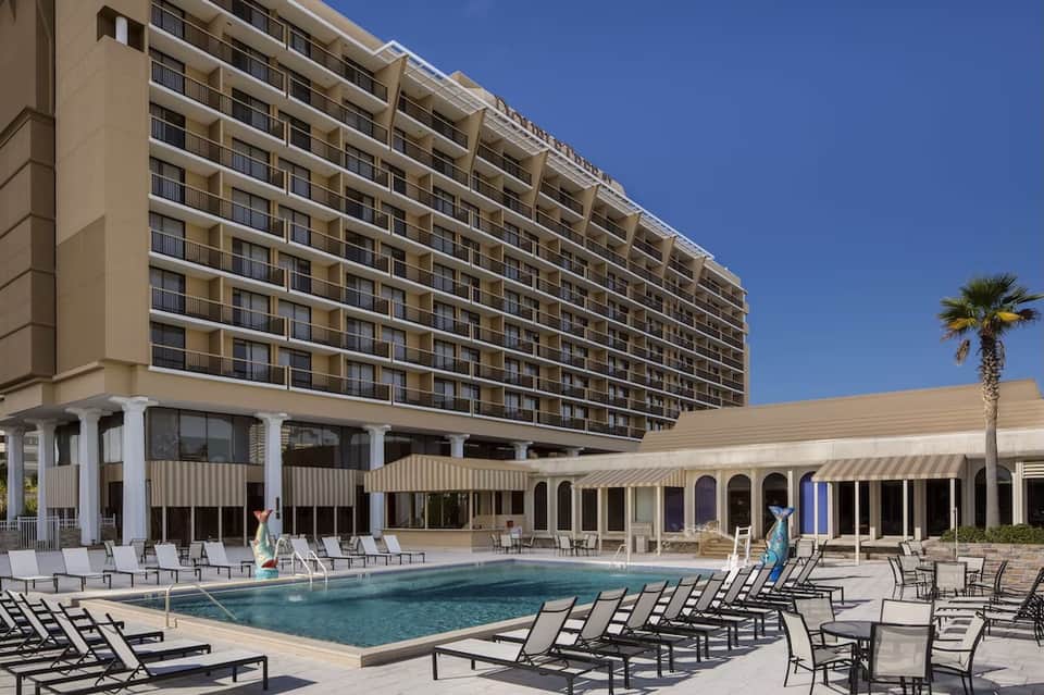 Resort pool and deck area with lounge chairs, umbrella covering, high-rise hotel building behind