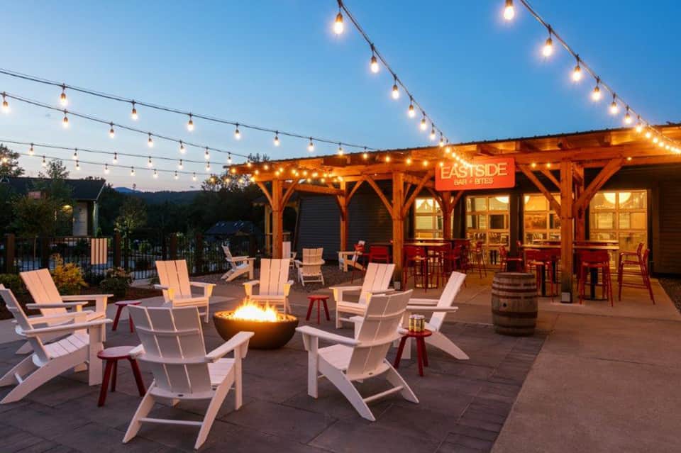 Evening outdoor patio with fire bowl, white Adirondack chairs, string lights, and wooden bar structure