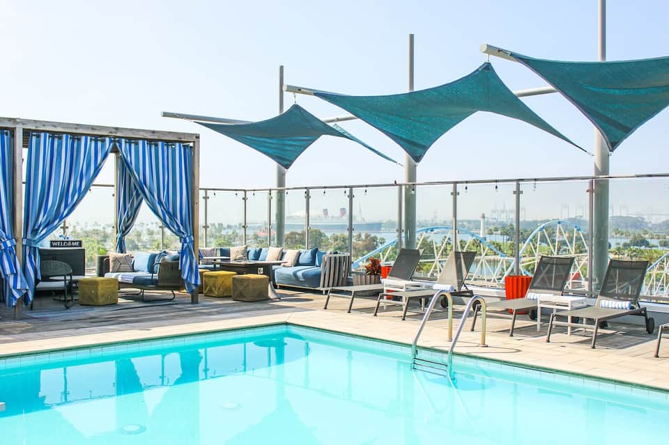Rooftop pool deck with teal shade sails, lounge seating, blue cabana, and city skyline view
