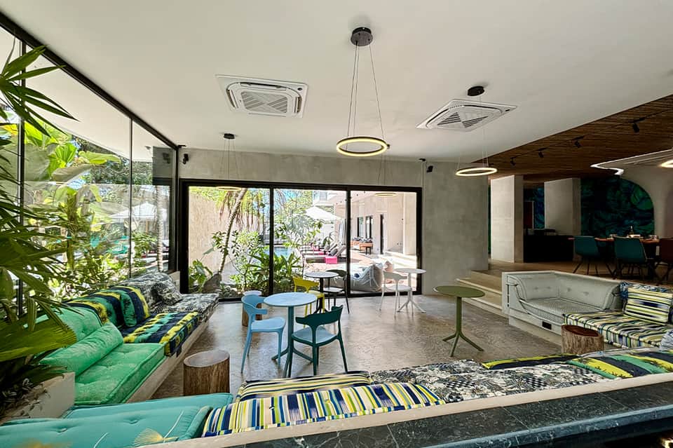 Modern resort lounge with green seating, indoor plants, concrete ceiling, pendant lights, and open courtyard views