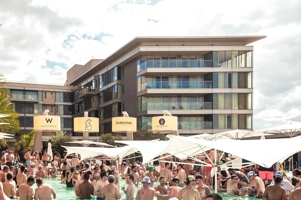 Daytime pool party with crowds swimming, white umbrellas, modern hotel building, and palm trees