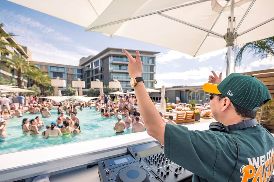 DJ at outdoor pool party with large crowd swimming and socializing under white umbrellas