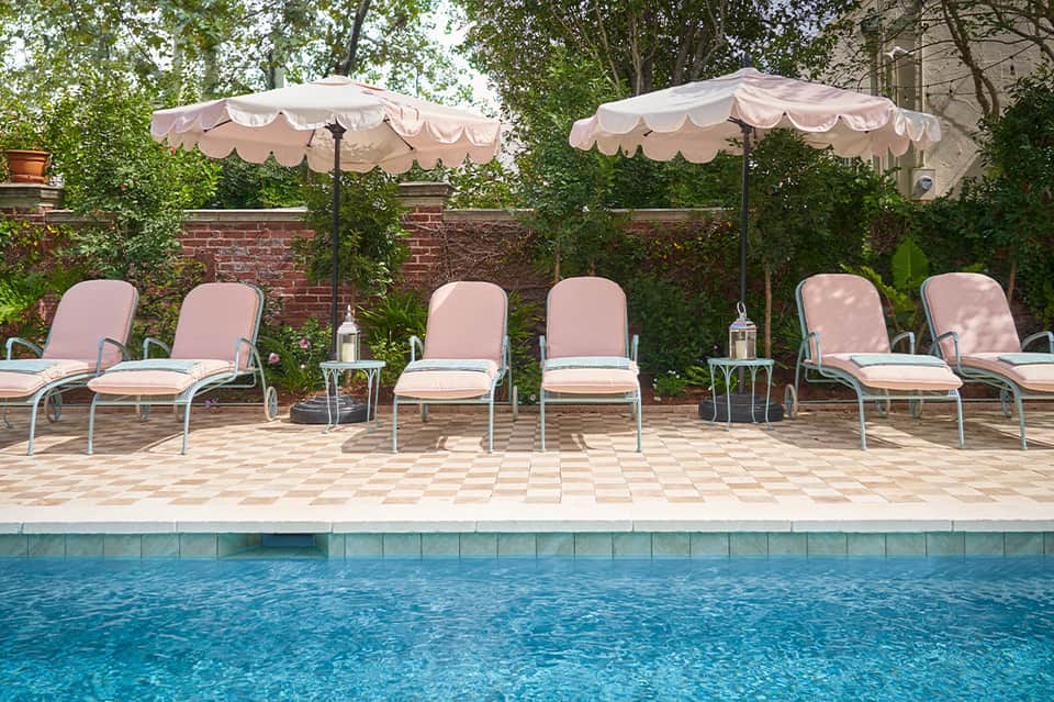 Poolside seating area with pink umbrellas, lounge chairs, brick wall, and evergreen landscaping