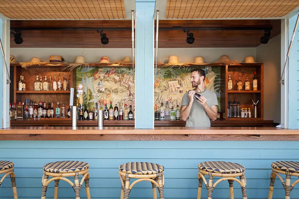 Beachside bar with wooden shelves of spirits, straw hats, and bartender in striped shirt