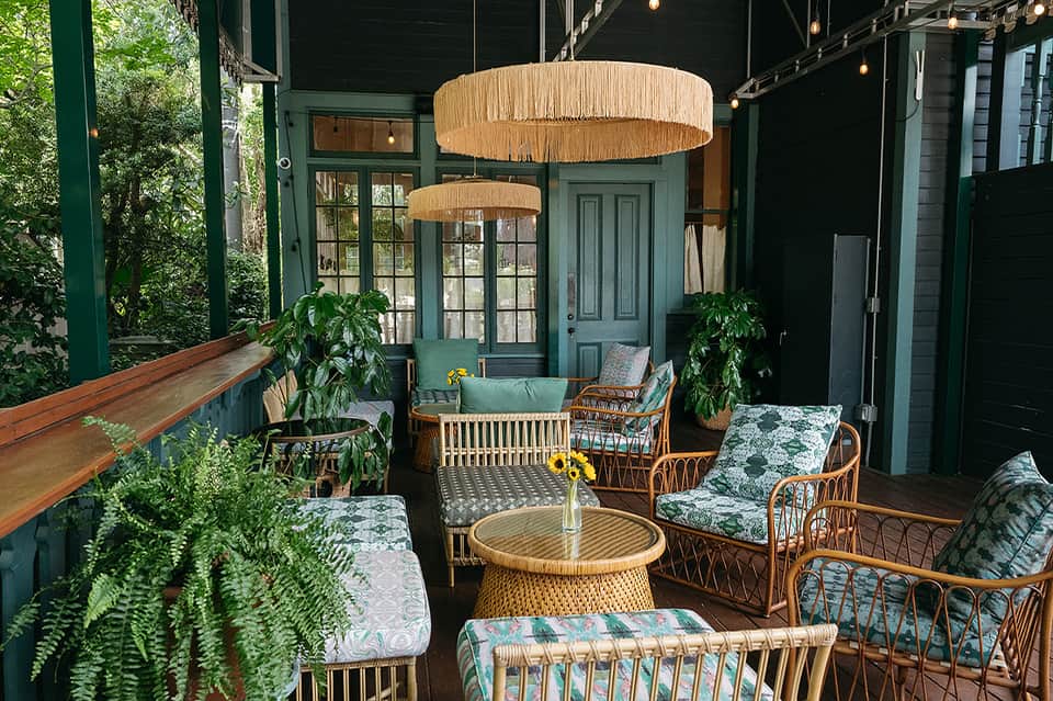 Green porch with wicker seating, rattan pendant lights, plants, and view of trees and garden area