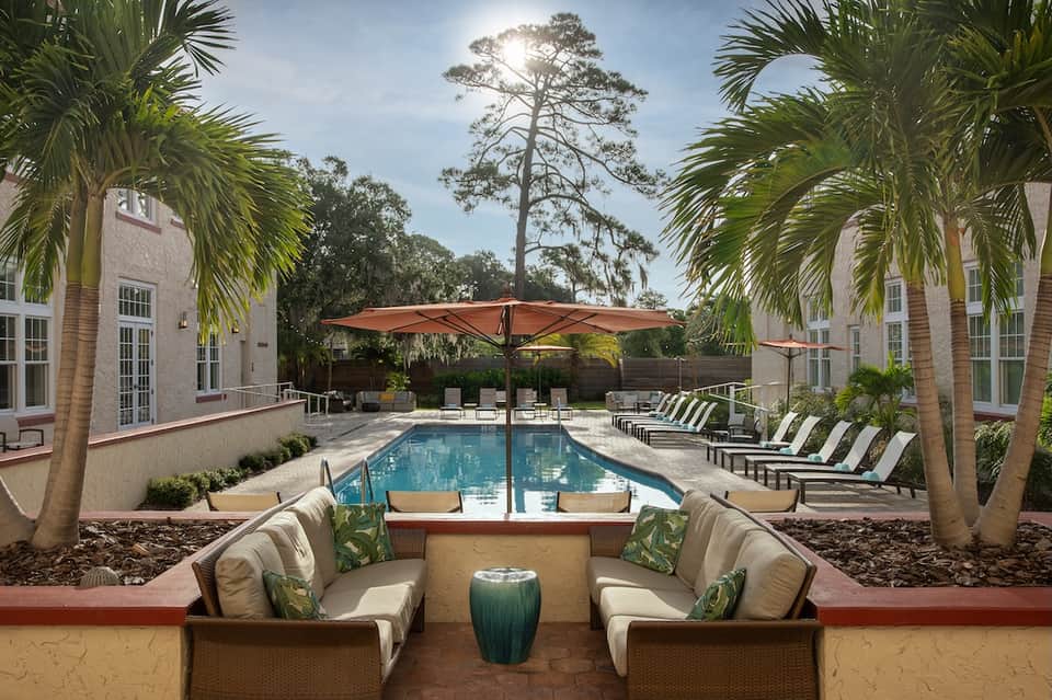Resort swimming pool lined with palm trees, umbrellas, and lounge chairs between modern buildings